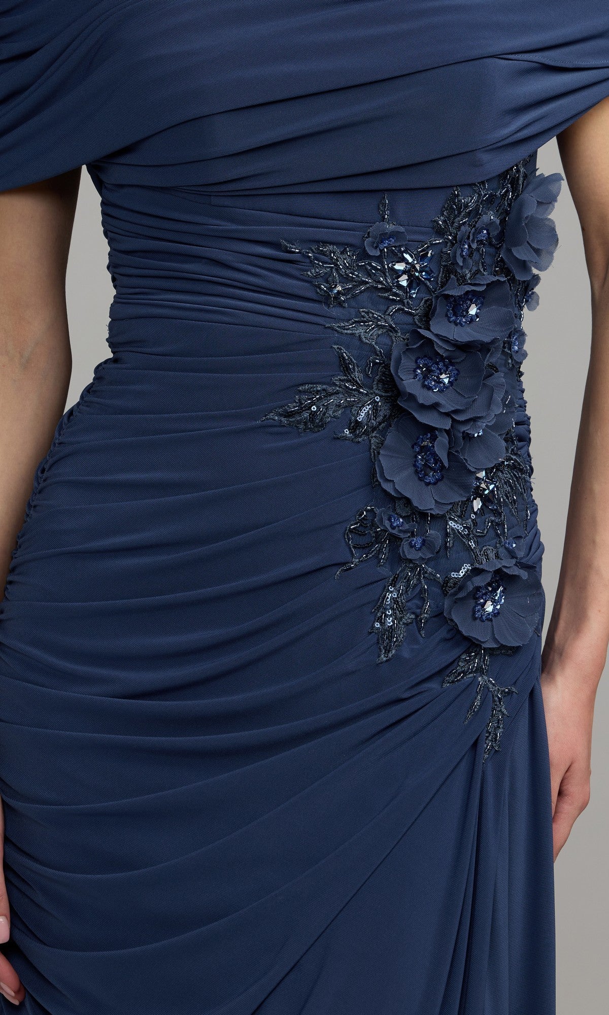 Close-up of the Long Formal Dress: Mac Duggal 13244 in navy blue, showcasing elegant ruching adorned with floral appliqué and beaded embellishments on the side—ideal for a sophisticated mother-of-the-bride ensemble. One hand is visible, drawing attention to the luxurious detailing.